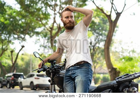 Concerned Tourist Travelling By Motorbike And Lost In City. Bearded Young Man Leaning On Motorcycle,