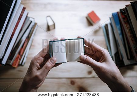 A Man Reading A Miniature Book Among The Big Books In The Library At The Wooden Table.