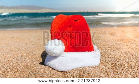 Closeup Photo Of Santa Clauss Hat Against Ea Waves On Beach At Sunny Day. Concept Of Travel And Tour