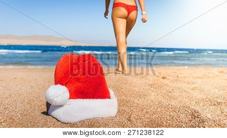 Closeup Photo Of Santa Clauss Hat Lying On The Beach Against Sexy Woman In Red Bikini. Concept Of Tr