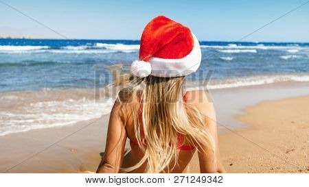 Sexy Young Woman With Long Hair Wearing Santa Clauss Hat Sitting On Beach At Sunny Windy Day. Concep
