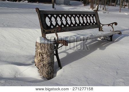 An Iron And Wooden Park Bench Alone And Covered By Snow With A Wood Log Used As A Side Table.