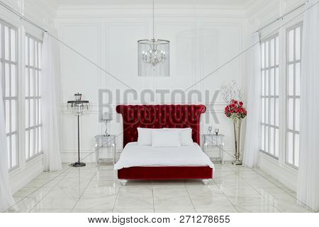 MOSCOW, RUSSIA - DEC 23, 2017: Interior of spacious white bedroom with large red bed, chandelier, lamps, flowers, windows on both sides in Ligtning Hall of Fotoelite Studio.