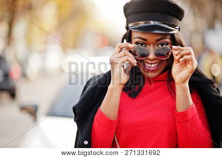 African American Fashion Girl In Coat, Newsboy Cap And Sunglasses Posed At Street Against White Busi