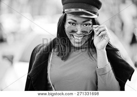 African American Fashion Girl In Coat, Newsboy Cap And Sunglasses Posed At Street Against White Busi