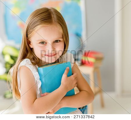 Little smiling blond girl standing near blackbord with red backpack and holding a notebook with pencils in the sunshine school classroom