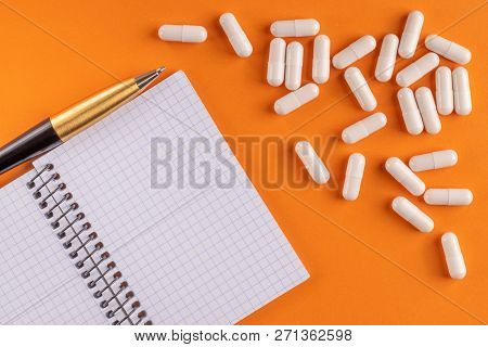 Medical Capsules And Pills Around A Blank Notebook With Pen On An Orange Background, Close-up, Top V