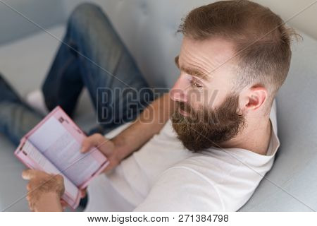 Pensive Frowning Handsome Man Reading Book At Home. Thoughtful Bearded Guy Sitting On Sofa And Looki