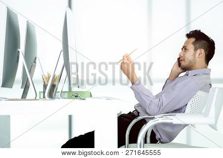 Young Asian Businessman Using Smartphone Sitting At Desk.  The Gesture Of Thinking In A Pencil Hand 