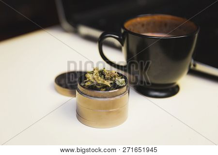 Cannabis Or Marijuana Flower In A Steel Grinder On A Desk, Next To A Cup Of Coffee And A Laptop.