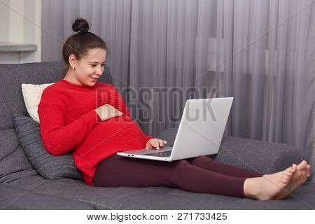 Indoor Shot Of Pleased Dark Haired Pregnant Young Woman Enjoys Domestic Atmosphhere, Keyborads Infor
