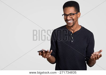 Happy Pleased Dark Skinned Guy Spreads Hands, Holds Cell Phone, Waits For Call Or Text Message, Smil
