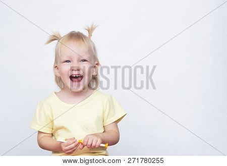Little Blue-eyed Blonde Girl Holding A Toothbrush In Her Hand And Laughing With An Open Mouth On A W