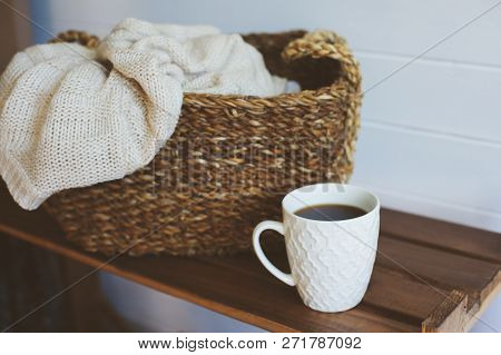 Cozy Winter Interior Details In White And Brown Tones. Basket With Knitted Sweater And Cup Of Coffee