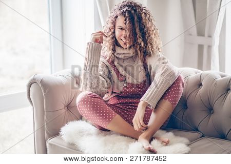 Hipster teenage girl with curly hair wearing beige knitted sweater and pink polka dot jumpsuit relaxing on a couch indoor