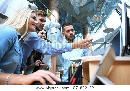 Group Of Young People In Casual Wear Sitting At The Office Desk And Discussing Something While Looki