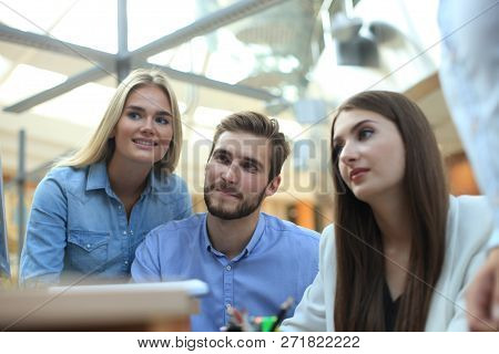 Group Of Young People In Casual Wear Sitting At The Office Desk And Discussing Something While Looki