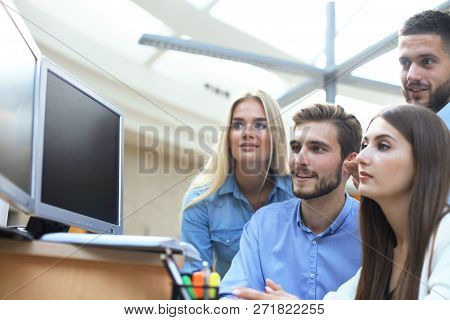 Group Of Young People In Casual Wear Sitting At The Office Desk And Discussing Something While Looki