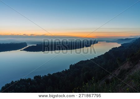 View From High Shore On River. Riverbank With Forest Under Thick Fog. Dawn Reflected In Water. Yello