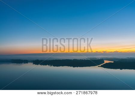 Broad River Flows Along Diagonal Shore With Forest Under Thick Fog. Early Blue Sky Reflected In Wate