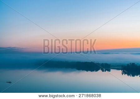 Broad River Flows Along Diagonal Shore With Silhouette Of Forest And Thick Fog. Tree Drifts With Flo