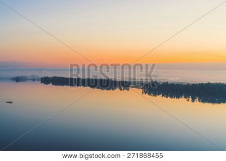 Broad Mystical River Flows Along Diagonal Shore With Silhouette Of Trees And Thick Fog. Gold Glow In