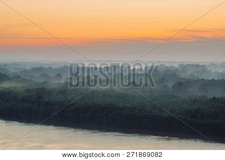 Mystical View On Riverbank Of Large Island With Forest Under Haze At Early Morning. Mist Among Layer