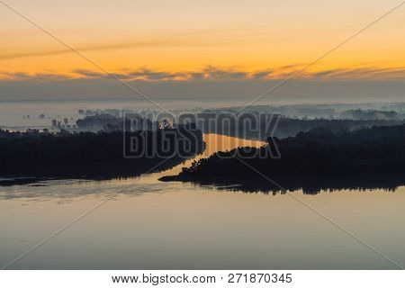 Riverbank With Forest Under Predawn Sky. Early Blue Sky Reflected In River Water. Yellow Stripe In P