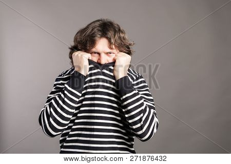 Young Man Pulling Black And White Striped Sweater Over Head Against Gray Background. Angry Male Pick