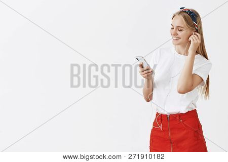 Studio Shot Of Joyful Modern Young Female In Red Skit And Headband Standing Half-turned While Puttin