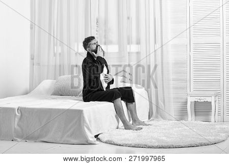 Awakening Concept. Man In Robe Sits On Bed, White Curtains On Background. Macho With Beard And Musta