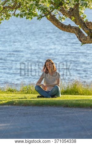 A Red-haired Girl Sits Under A Flowering Tree By The Lake And Enjoys The Sunset, Girl Looking At The