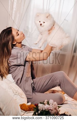 A Girl In Pajamas Sits In Bed At Night With Her White Dog Watching A Laptop And Eating Sweets.girl W