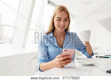 Smiling Young Woman With Cellphone In Hand While Drinking Coffee In A White Room