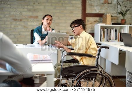 Portrait Of Young Male Office Worker In A Wheelchair Talking To Female Colleague While Showing Her I