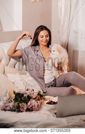 A Girl In Pajamas Sits In Bed At Night With Her White Dog Watching A Laptop And Eating Sweets.girl W