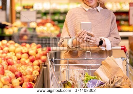 Mature female consumer in knitted beige sweater pushing cart with food products in supermarker and scrolling in smartphone