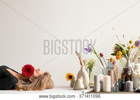 Young serene woman with red herbera in her mouth lying by variety of fresh and dried flowers and wildflowers in vases and group of candles