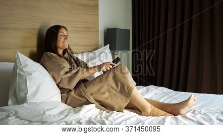 Smiling Lady In Beige Bathrobe With Remote Control In Hand Watches Tv At Coronavirus Self-isolation 