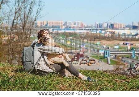 A Girl Sits Hugging A Labrador Retriever On A Mountain Against The Background Of The City. Vacation 