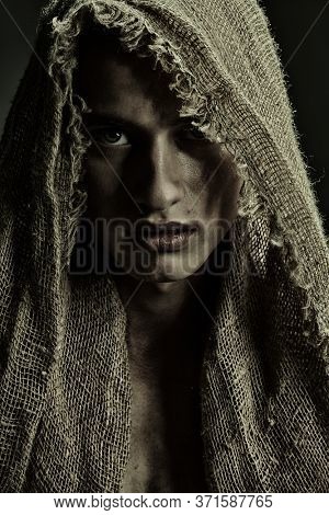 Portrait of a handsome young man in burlap clothes on a black background. Medieval wanderer, pilgrim.