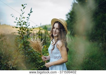 Smiling Girl In A Straw Hat Collects Flowers. Girl In A Summer Dress In The Tall Grass. Woman In A G