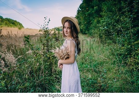 Happy Young Woman In A White Dress In A Straw Hat. Smiling Girl With A Bouquet Of Dried Flowers. Gir
