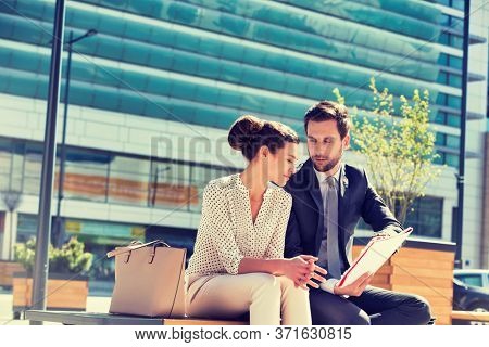 Young attractive businessman discussing business plans over document with beautiful businesswoman while sitting on bench against office building
