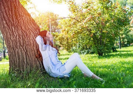 Young Woman Sitting Under Tree In Summer Park On Sunny Day. Beautiful Redhead Girl Relaxing With Clo