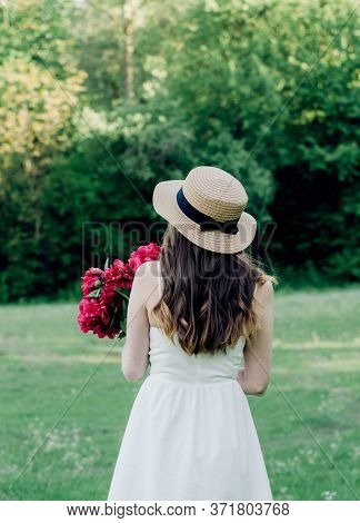 Girl With Long Hair In A Boater Hat Stands With Her Back In A White Dress And Holds A Bouquet Of Red