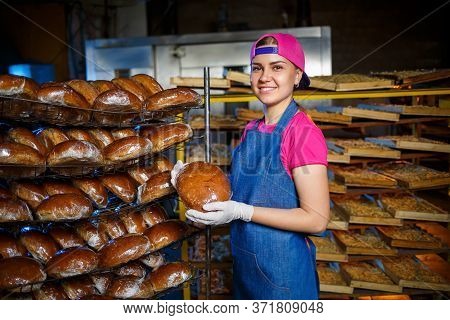 Smiling Baker Girl In Apron Holding Bread And Looking At Camera. Young Woman Is A Chef Holding Fresh