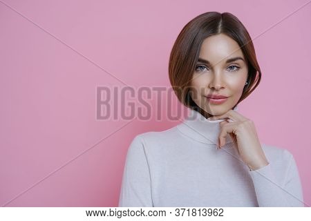 Portrait Of Brunette Woman Keeps Hand On Collar Of Turtleneck, Has Healthy Perfect Skin, Wears Minim