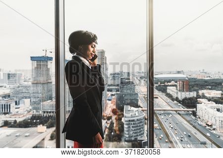 A Serious African-american Businesswoman Is Talking On The Phone While Standing Near A Window, On A 