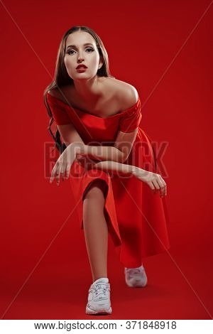 Beautiful brunette girl posing at studio in red dress and white sneakers. Red background. Sport chic style.